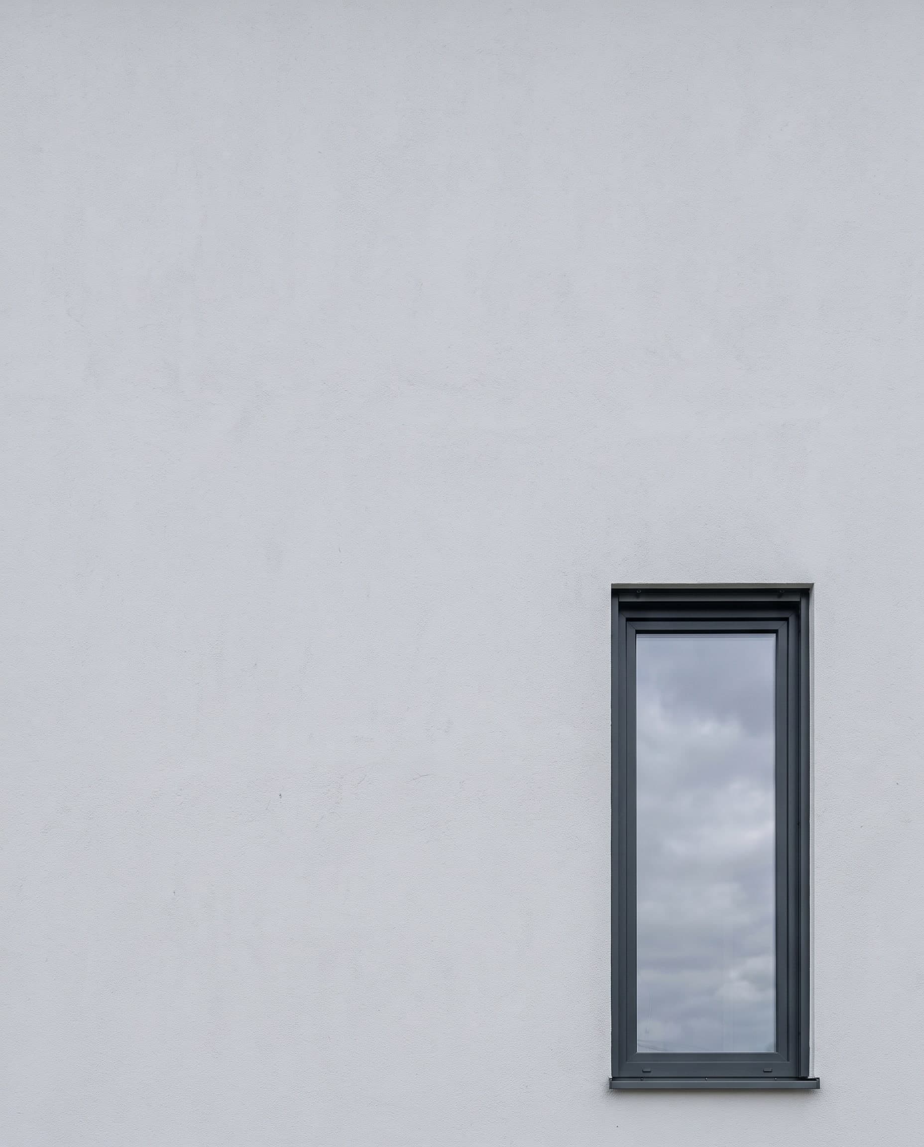 Hell verputzte Hauswand mit einem hohen Fenster in dunklem Rahmen, in der Scheibe spiegelt sich der Himmel